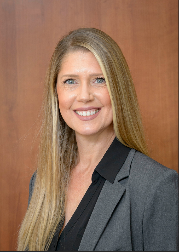 Tilly Clark Professional headshot of Tilly Clark, Director of Presidential Affairs and Event Planning at the University of Texas Medical Branch, wearing a gray blazer and dark blouse, photographed against a warm wood‑tone background.