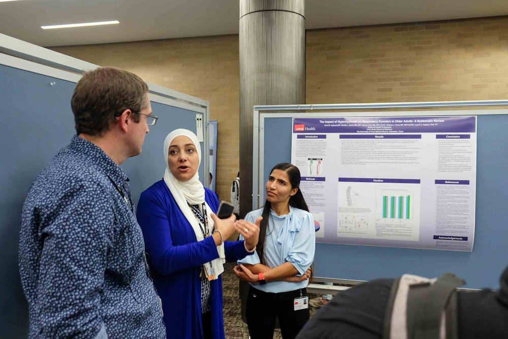 A student speaks with two attendees in front of a research poster during a graduate research discussion.
